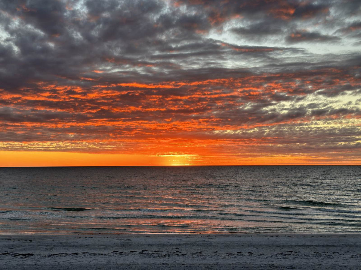 Sunset over the beach, with the sun touching the horizon under orange and gray clouds