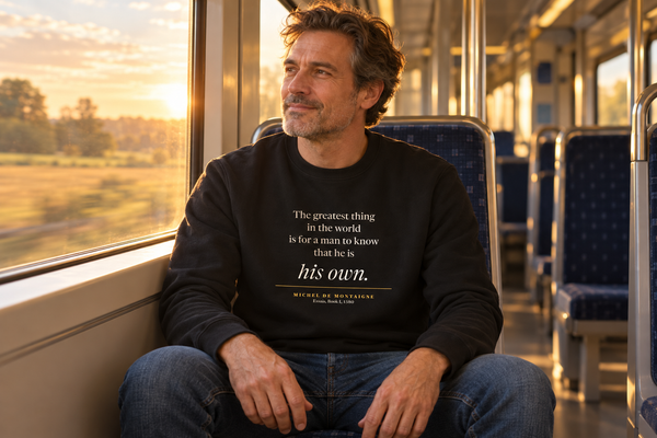 Man on a train at golden hour wearing The Greatest Thing in the World — Montaigne sweatshirt.